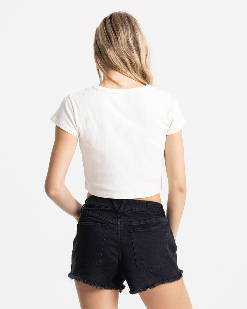 A woman with long blonde hair, seen from behind, wears the Women’s Ride Teehe Wave 1991 Mid Tee by Volcom and high-waisted black denim shorts against a plain white background.