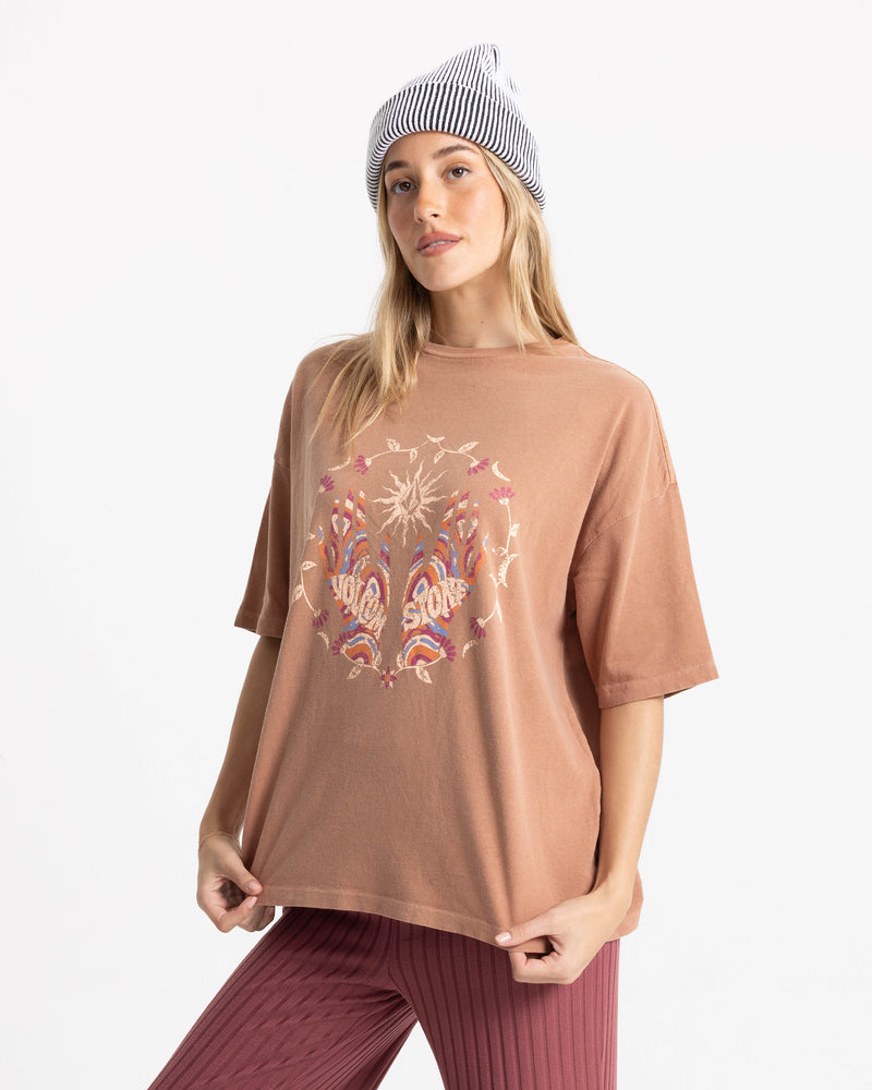 A woman wears the Volcom Women’s Mystic Palms Bad Tee, maroon ribbed pants, and a striped beanie, standing against a white background in a relaxed pose.