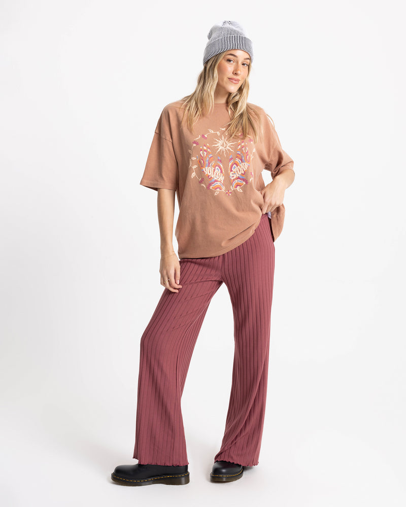 A woman in a gray beanie, loose Women’s Mystic Palms Bad Tee by Volcom, and pink pleated pants stands against a white background, one hand in her pocket and wearing black shoes.
