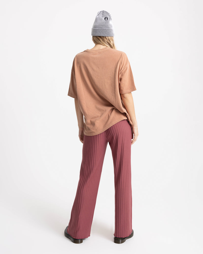 A person faces away, wearing the Volcom Women’s Mystic Palms Bad Tee in beige, pink pleated pants, black shoes, and a gray striped beanie against a plain white background.