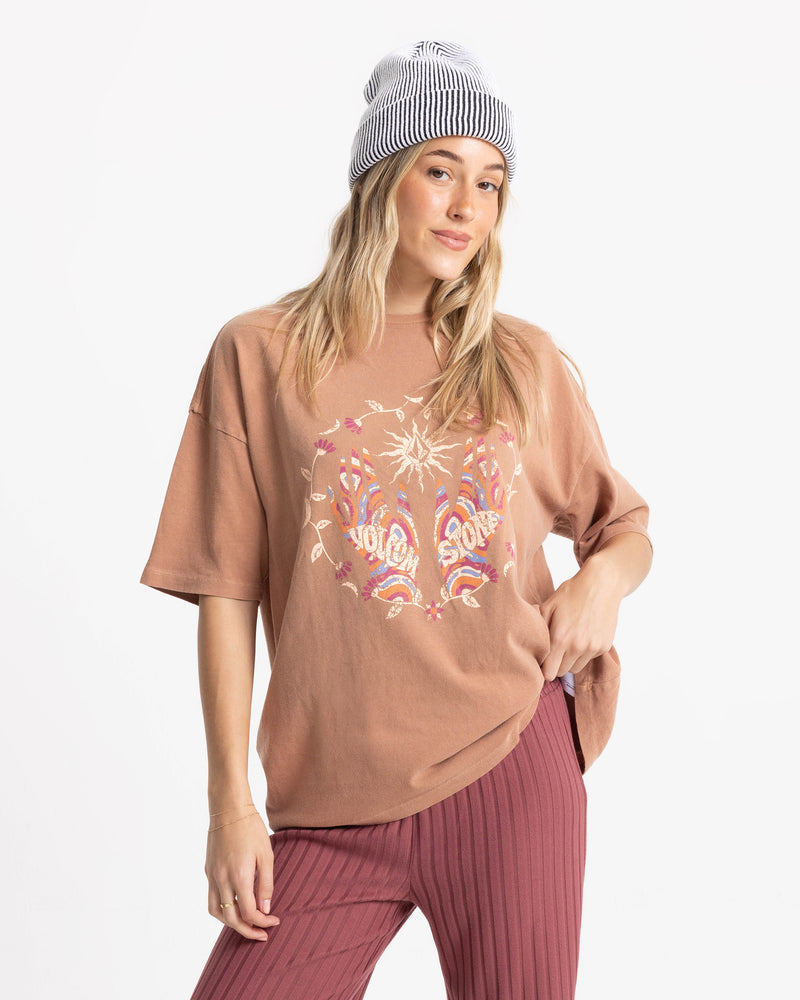A woman in a striped beanie and mauve pants smiles slightly against a white background, wearing the Volcom Women’s Mystic Palms Bad Tee.