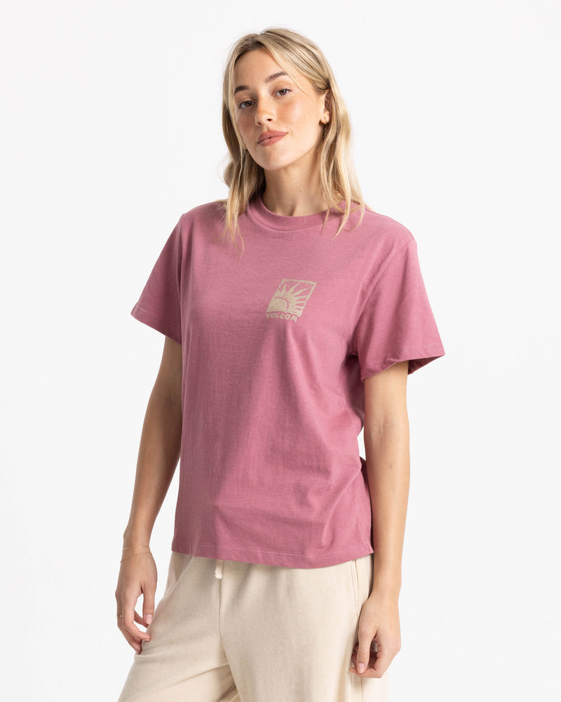 A person wears a dusty rose Women’s Volcom Stamp Lock It Up Tee in a relaxed, boxy fit, featuring a small square print of a sun rising over a horizon on the left chest.