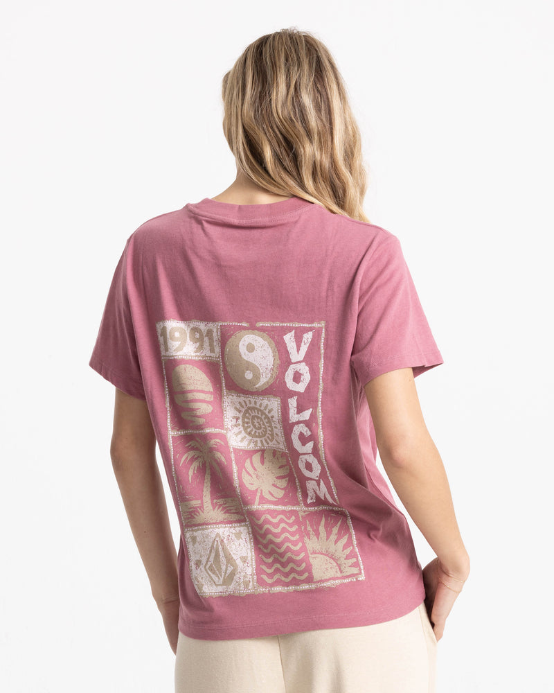 A person seen from behind wearing a dusty rose Women’s Volcom Stamp Lock It Up Tee, featuring a back graphic grid with a yin-yang symbol, a palm tree, and the text 1991.