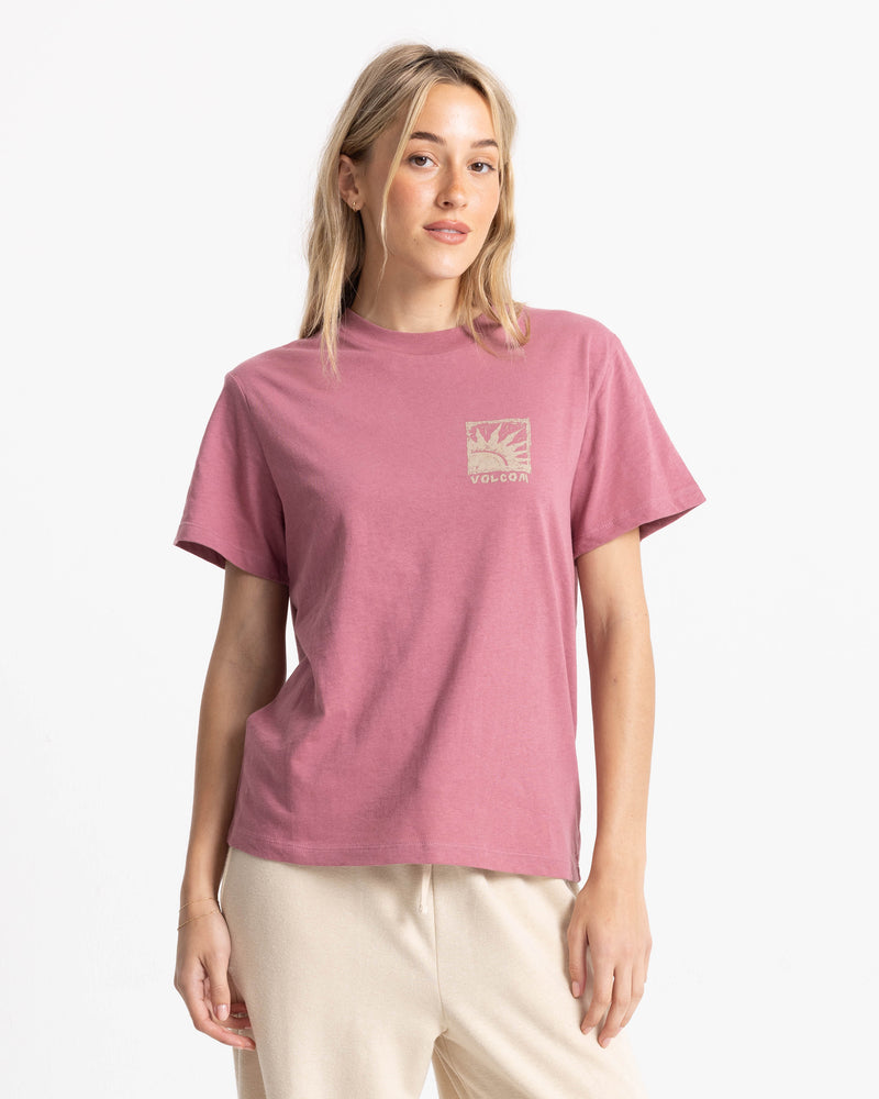 A woman wearing a mauve, relaxed boxy fit Women’s Volcom Stamp Lock It Up Tee with a small square graphic on the left chest depicting a sun and the word Volcom.