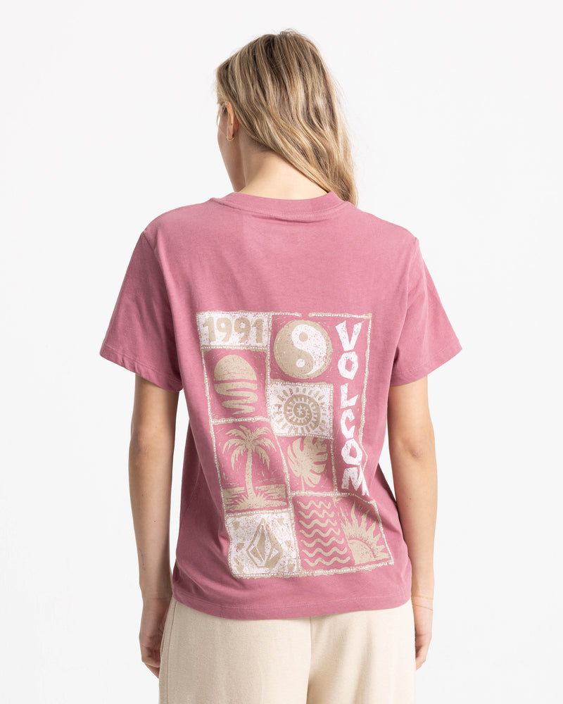 Back view of a person wearing a dusty pink, relaxed boxy fit Women’s Volcom Stamp Lock It Up Tee, featuring a 1991 graphic with a yin-yang symbol, a palm tree, and assorted geometric designs.