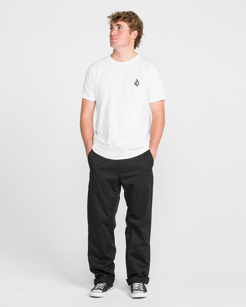 A person stands against a plain white background wearing Men's Frickin Relaxed Chino Pants, a white short-sleeve t-shirt with a small chest logo, black-and-white sneakers, hands in pockets, and looking upward.