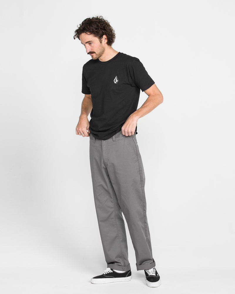 A man with curly hair and a mustache stands against a plain white background, wearing Men's Frickin Relaxed Chino Pants in gray, a black T-shirt with a small white logo, and black sneakers.