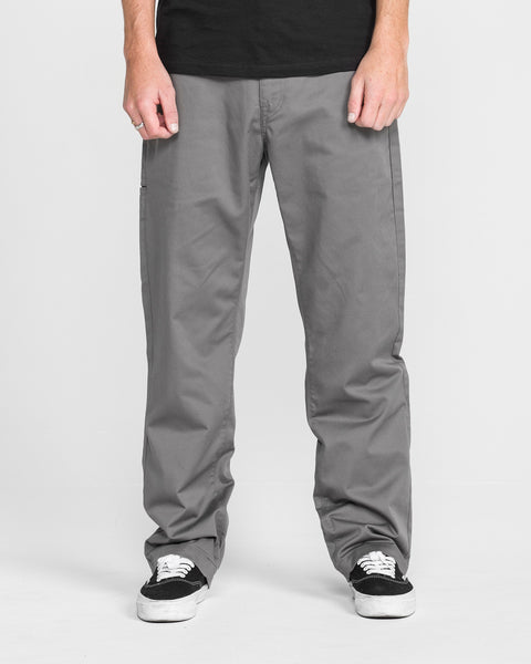 A person wears Men's Frickin Relaxed Chino Pants with black-and-white sneakers, standing against a plain white background. Their hands rest at their sides, and the photo is cropped from the shoulders down.