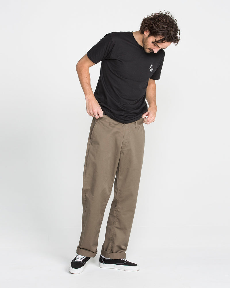 A man with curly hair wearing a black t-shirt, black skate shoes, and Men's Frickin Relaxed Chino Pants stands against a plain white background, looking down and holding his pants at the waist.