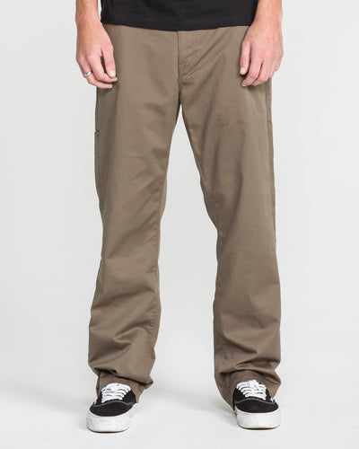 A person models the Men's Frickin Relaxed Chino Pants in khaki, paired with black-and-white sneakers, standing against a plain white background with only their lower torso, legs, and hands visible.