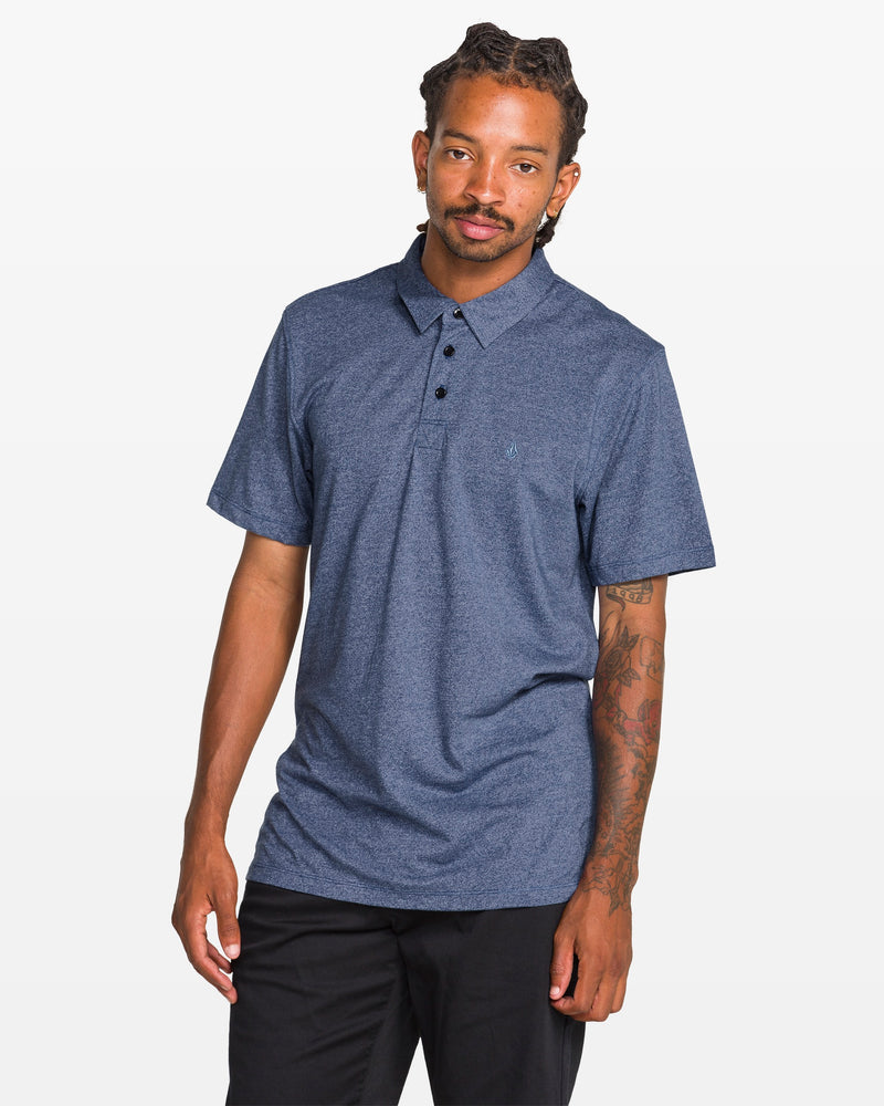 A man with dreadlocks and a tattooed left arm models the Volcom Men's Wowzer Polo Short Sleeve Shirt in blue, styled with black pants, standing against a plain white background.