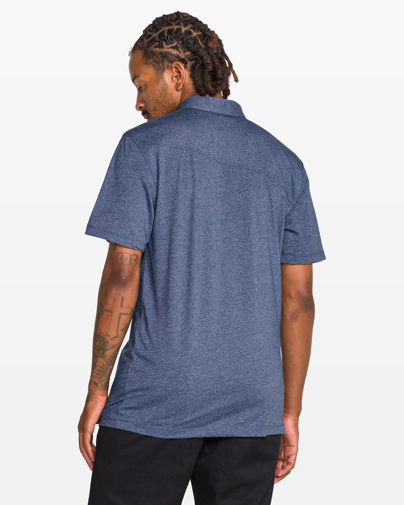 A person with short dreadlocks and visible right arm tattoos wears the Volcom Men's Wowzer Polo Short Sleeve Shirt in blue and black pants, standing against a plain white background and looking over their left shoulder.