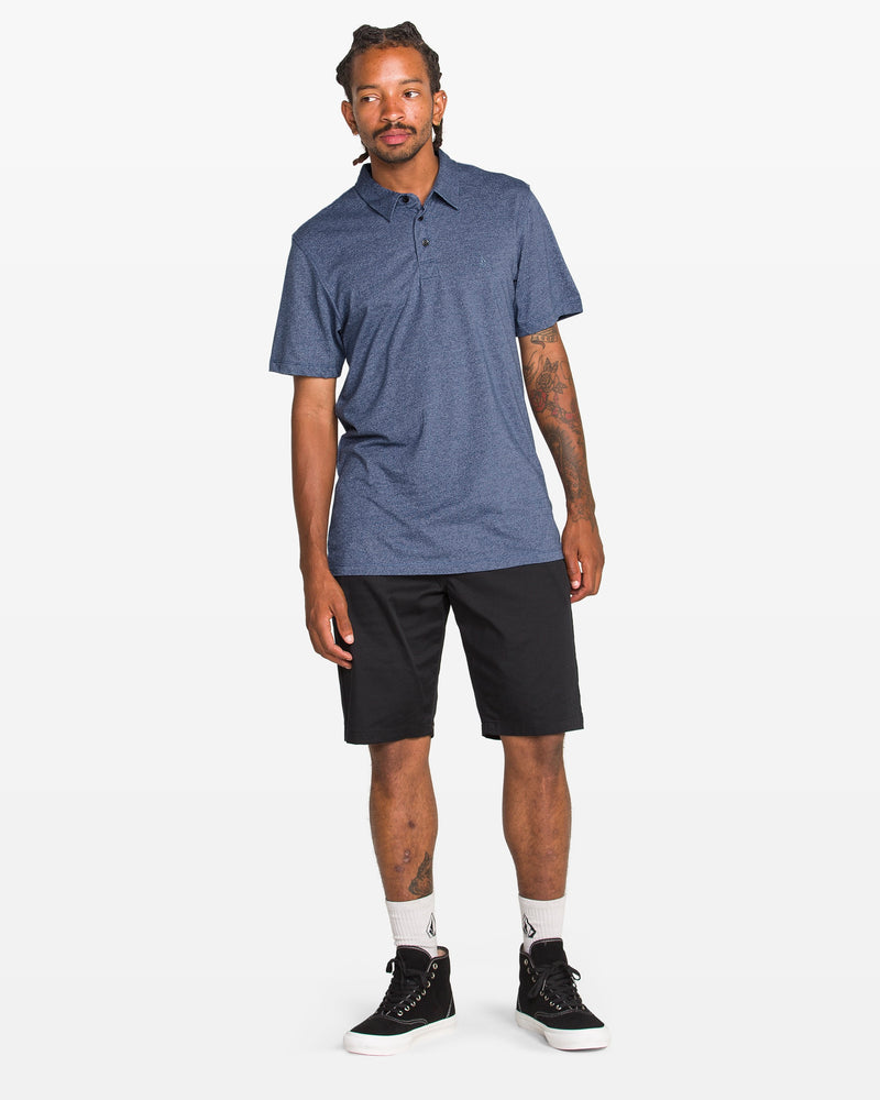 A man with short, braided hair and a neutral expression wears a blue Volcom Men's Wowzer Polo Short Sleeve Shirt, black knee-length shorts, white crew socks, and black sneakers against a plain white background.