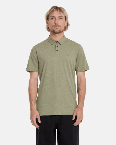 A man with light hair and a short beard wears the Volcom Men's Wowzer Polo Short Sleeve Shirt in light olive green and black pants, standing against a plain light background.