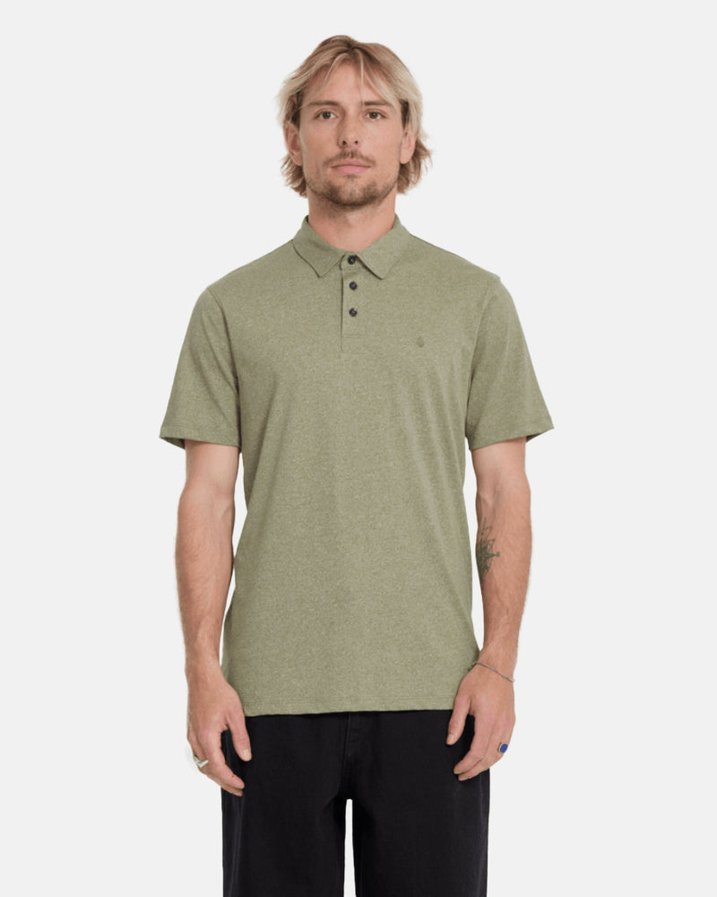 A man with light hair and a short beard wears the Volcom Men's Wowzer Polo Short Sleeve Shirt in light olive green and black pants, standing against a plain light background.