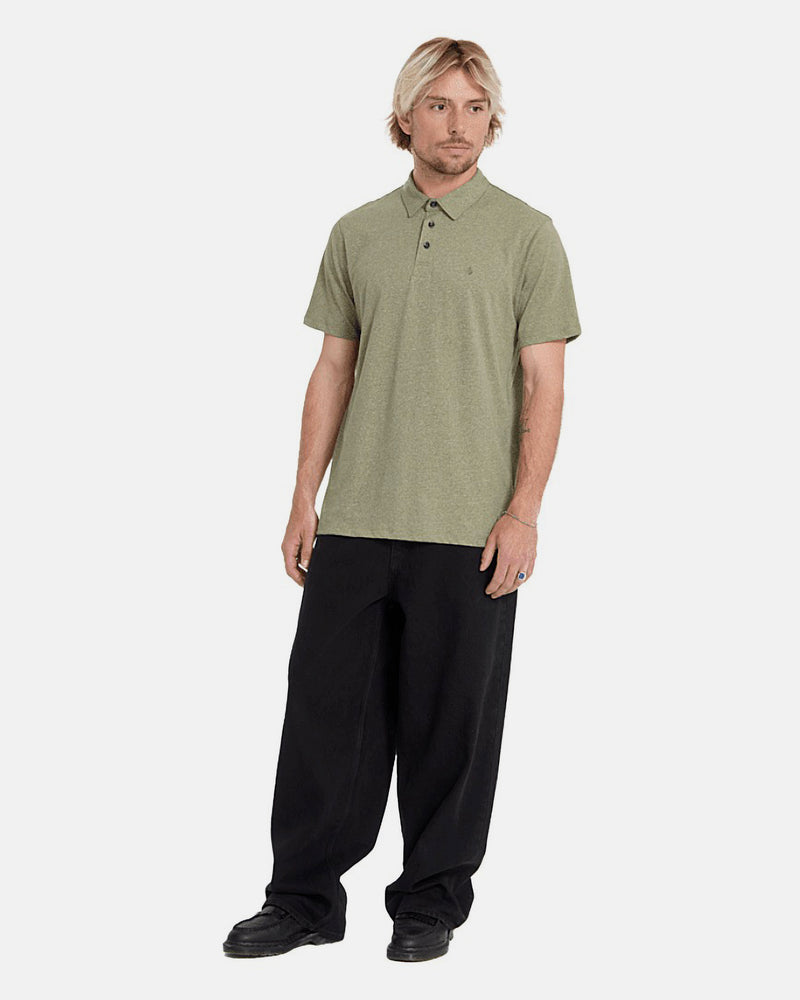 A man with blonde hair wears a Volcom Men's Wowzer Polo Short Sleeve Shirt in olive green, paired with loose black pants and black shoes. He stands against a plain white background, looking slightly to the side with a neutral expression.