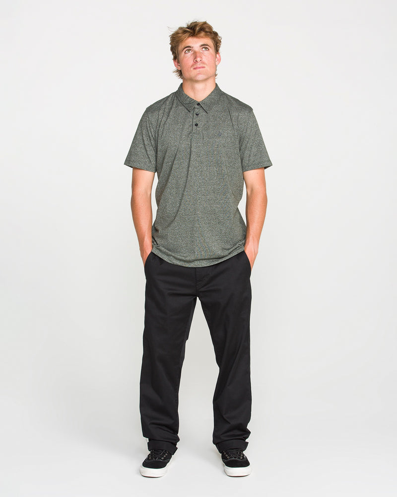 A man wearing the Volcom Men's Wowzer Polo Short Sleeve Shirt in gray, paired with black pants and black sneakers, stands with his hands in his pockets, looking upward against a plain white background.