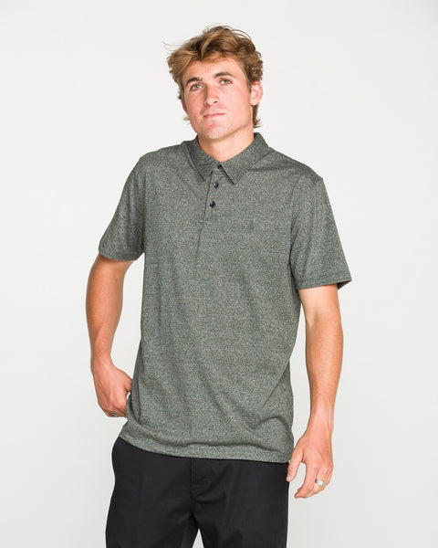 A young man with light brown hair wearing the Volcom Men's Wowzer Polo Short Sleeve Shirt in dark grey and black pants stands against a plain white background.