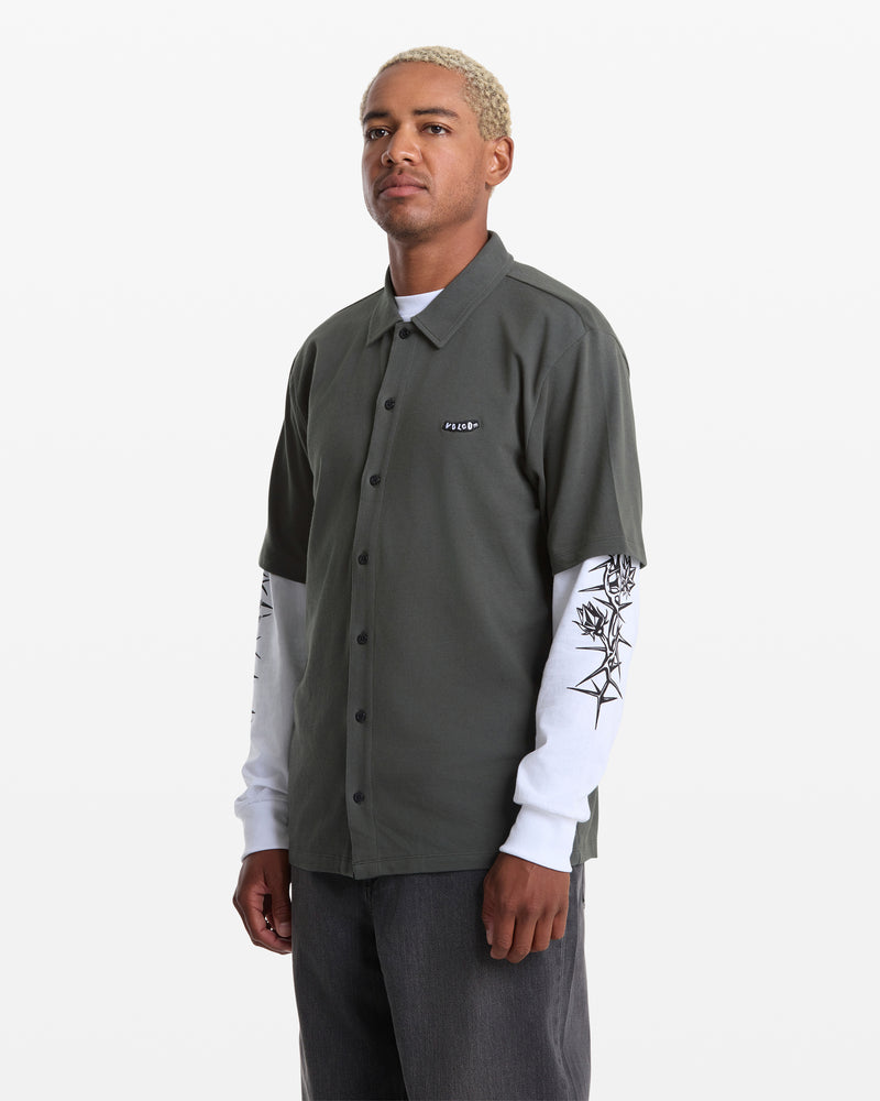 A person with short, bleached hair wears a dark green Volcom Men's Chavo Button Down Short Sleeve shirt over a white long-sleeve with black sleeve designs and dark pants, standing against a plain white background.