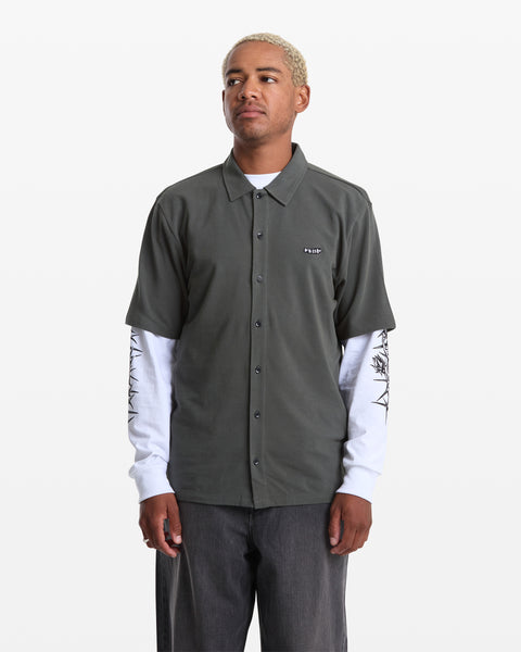 A man with short blond hair wears the Volcom Men's Chavo Button Down Short Sleeve over a white long-sleeve graphic shirt and dark pants, standing against a plain white background.
