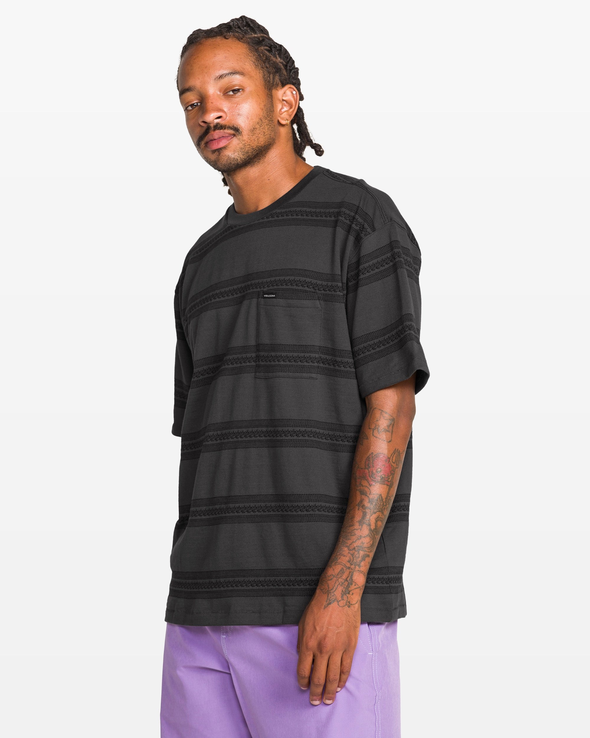 A man with braided hair and arm tattoos wears the Volcom Men's Yewster Crew, a loose-fitting black jacquard stripe short sleeve 100% cotton tee with a pocket, paired with light purple pants, standing against a plain white background.