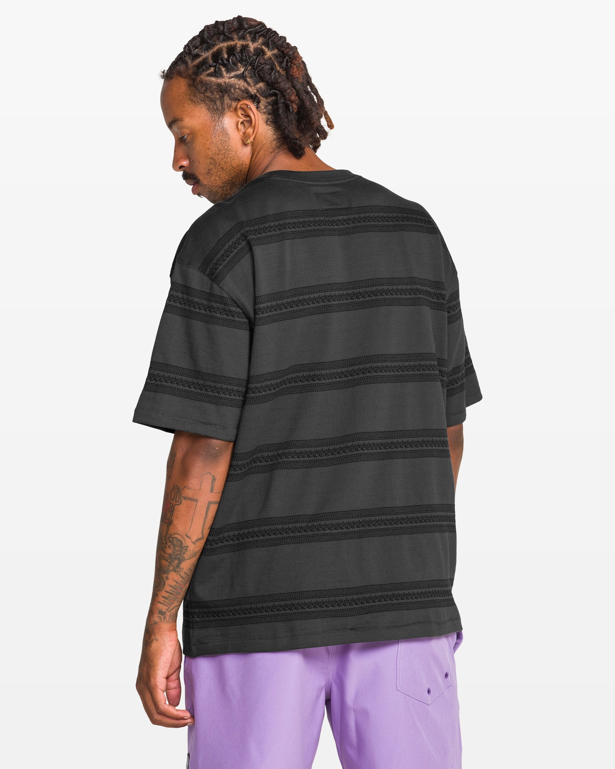 A man with dreadlocks, seen from behind, wears a Volcom Men's Yewster Crew black jacquard stripe tee and lavender shorts. He turns his head slightly, showing a tattoo on his left forearm. The background is plain white.