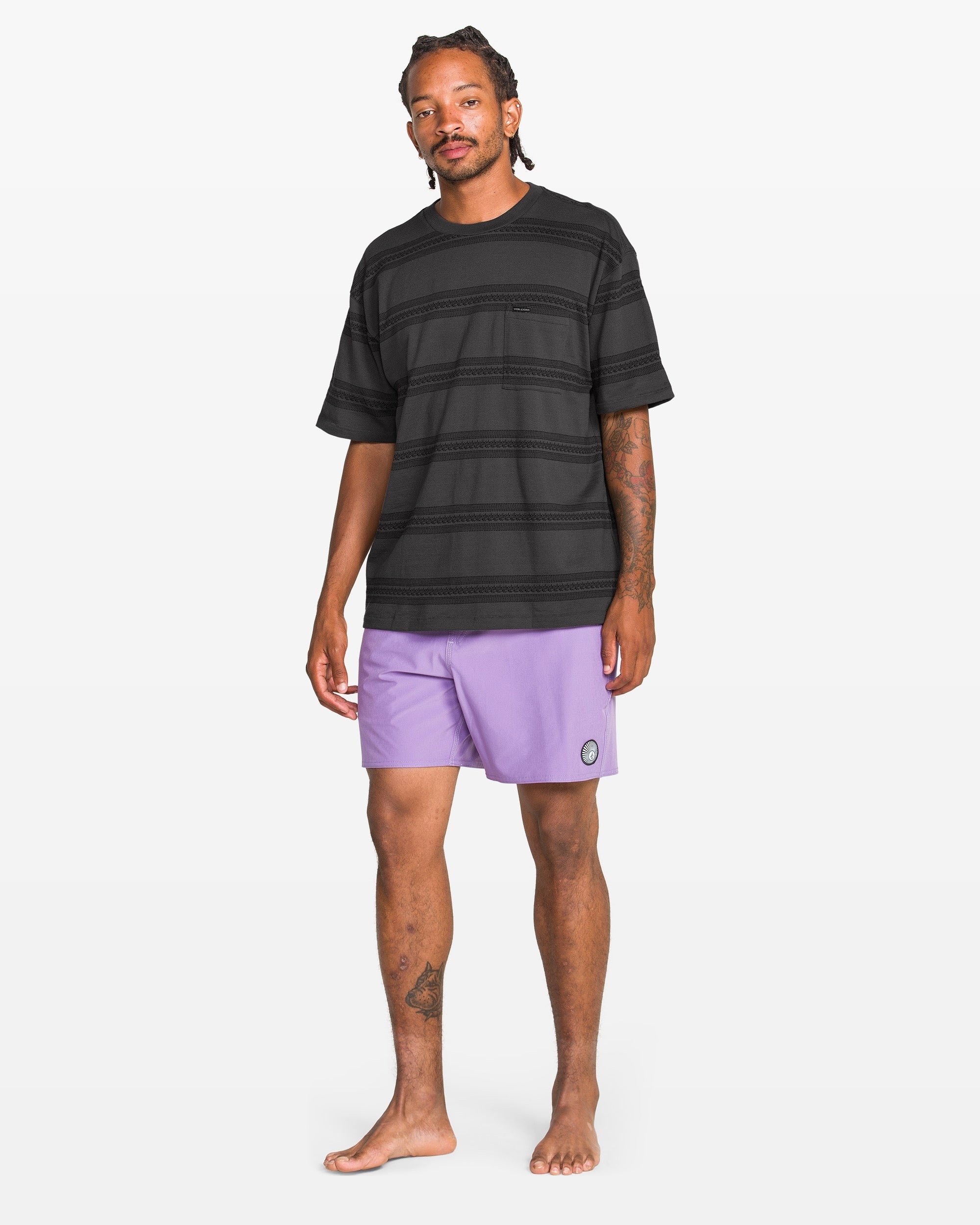 A man stands barefoot in a Volcom Men's Yewster Crew black patterned T-shirt and light purple shorts with a small patch on the left leg, against a plain white background.