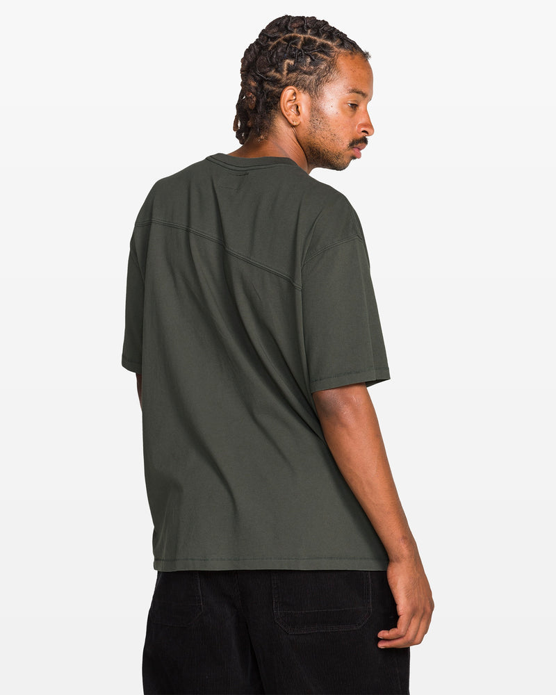 A person with braided hair stands sideways, looking over their shoulder, wearing a loose-fitting Volcom Men's Stone Cruiser Crew T-shirt in 100% cotton jersey (dark green) and black pants against a plain white background.