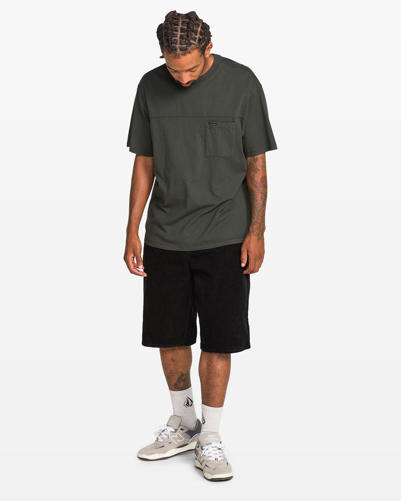 A man faces slightly downward, wearing a dark green Volcom Men's Stone Cruiser Crew tee with a chest pocket, black shorts, white socks, and light grey sneakers against a plain white background.
