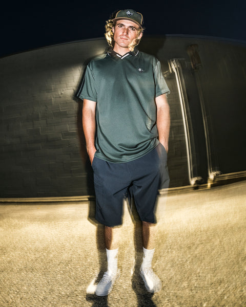 Wearing the Volcom Men’s Chillstone Short Sleeve Polo in green, along with navy shorts, a cap, white socks, and sneakers, a person stands outside at night against a blurred background with a lighting effect.