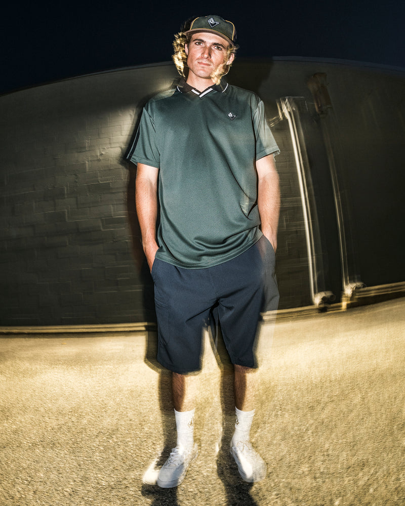 Wearing the Volcom Men’s Chillstone Short Sleeve Polo in green, along with navy shorts, a cap, white socks, and sneakers, a person stands outside at night against a blurred background with a lighting effect.