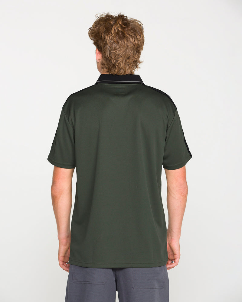 A person with light brown hair stands facing away, wearing the Volcom Men's Chillstone Short Sleeve Polo in dark green with a black collar and shoulders featuring subtle yarn-dye stripes, paired with gray shorts, against a plain white background.