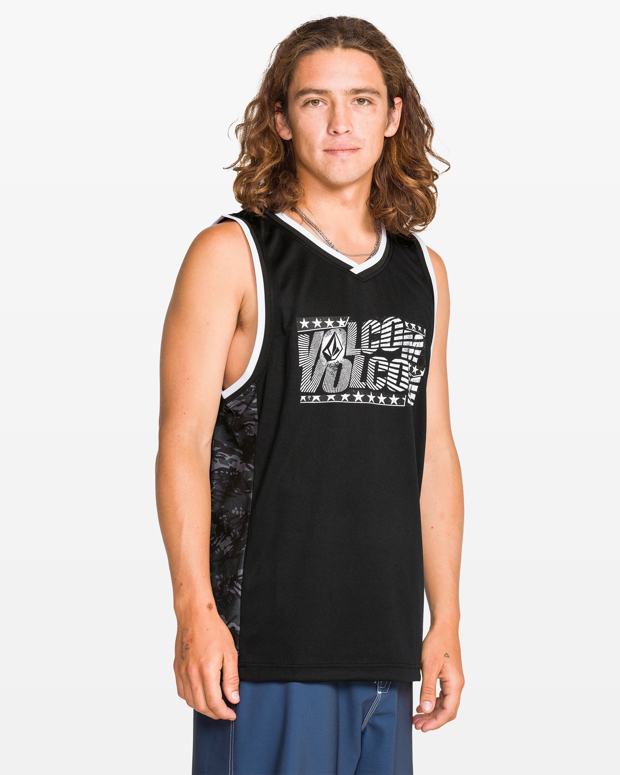 A young man with long brown hair wears a Volcom Men's Froth Of July Tank in black with white trim and a front graphic logo, paired with blue shorts, standing against a plain light background.