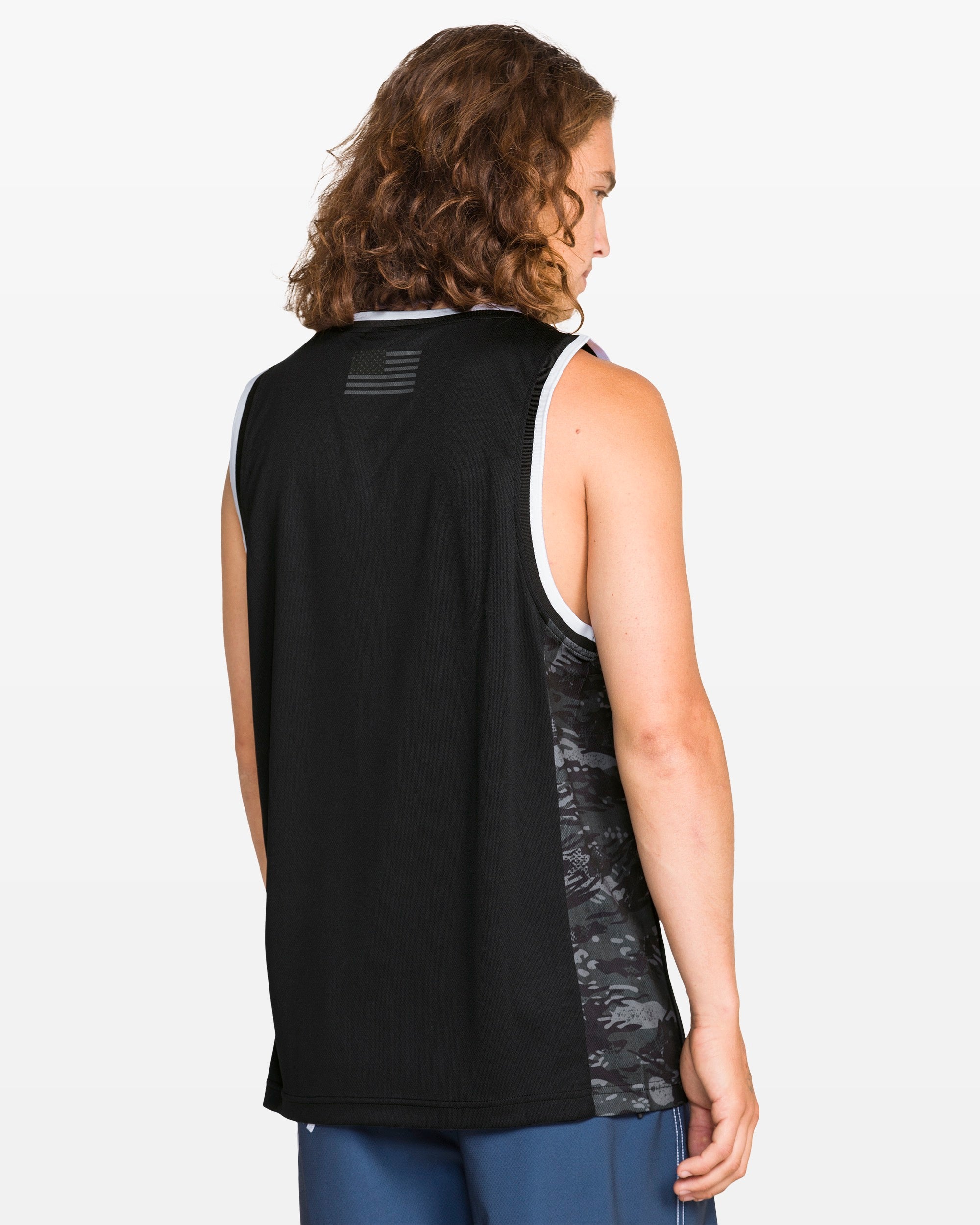 A person with long, wavy brown hair is seen from behind, wearing the Volcom Men's Froth Of July Tank and blue shorts, standing against a plain white background.