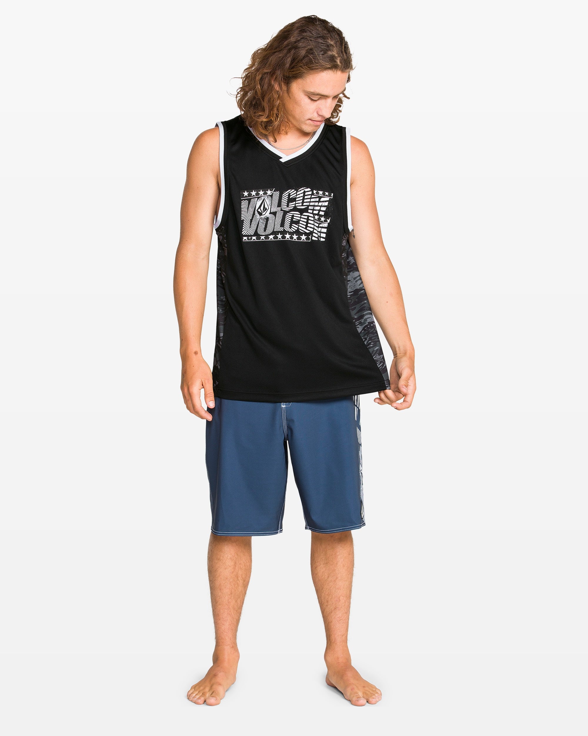 A barefoot person with long hair wears a black Volcom Men's Froth Of July Tank with a white logo and blue athletic shorts, standing and looking down at their side against a plain white background.
