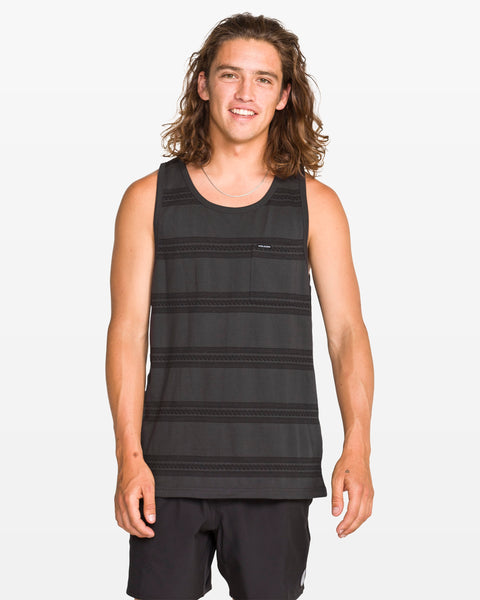 A young man with long brown hair stands and smiles at the camera against a plain white background, wearing a modern fit Men's Yewster Tank by Volcom in black and gray jacquard stripes paired with black shorts.