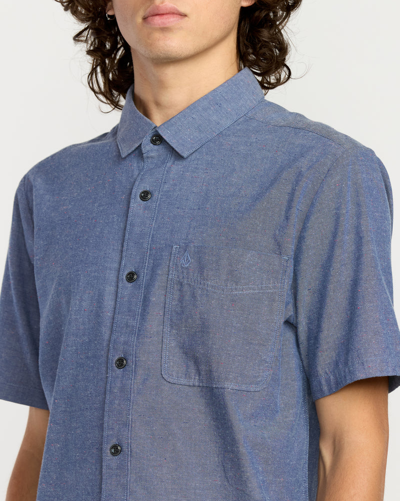 A person with long curly hair models the Men's Date Knight Short Sleeve Shirt, a classic-fit Volcom top in a premium cotton blend with a chest pocket and black buttons, against a plain light background. Their face is partially out of frame.