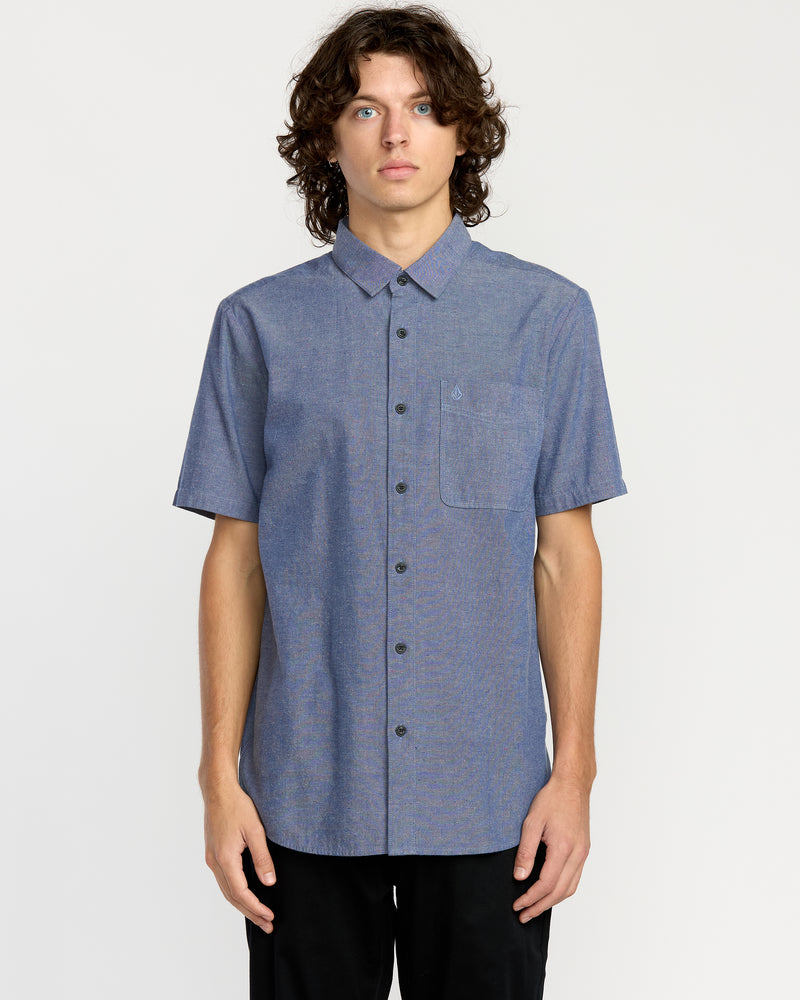 A young man with curly brown hair wears the Men's Date Knight Short Sleeve Shirt by Volcom, a classic-fit light blue shirt with a chest pocket, crafted from a premium cotton blend, standing against a plain white background.
