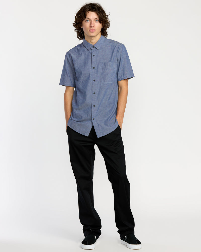 A young person with curly brown hair wears the Men's Date Knight Short Sleeve Shirt from Volcom—classic-fit, blue button-up style—paired with black pants and black slip-on shoes, standing against a plain white background with hands in pockets.
