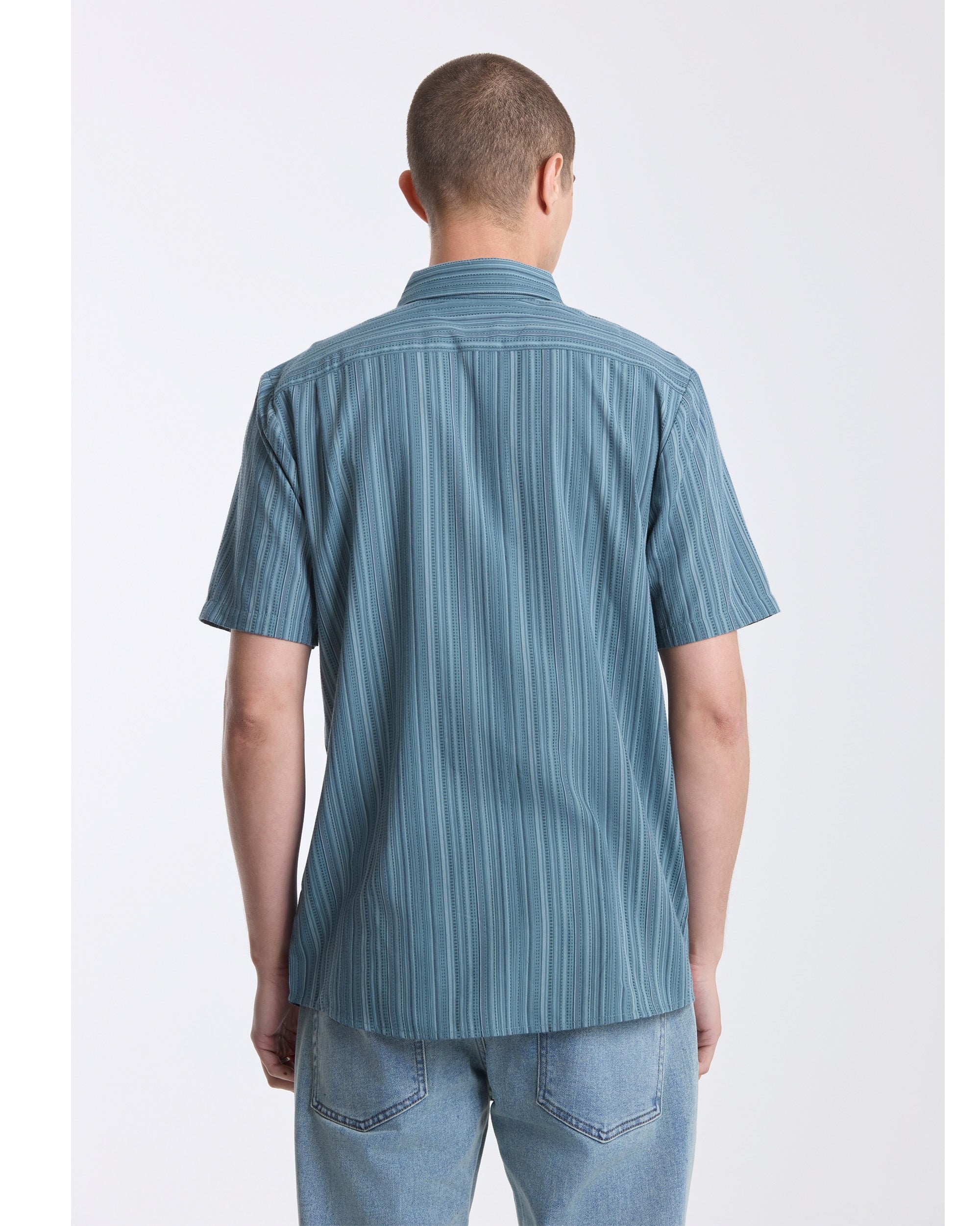 A person with short hair is seen from behind, wearing a blue Volcom Men's Jahstripe Short Sleeve Shirt and light blue jeans against a plain white background.