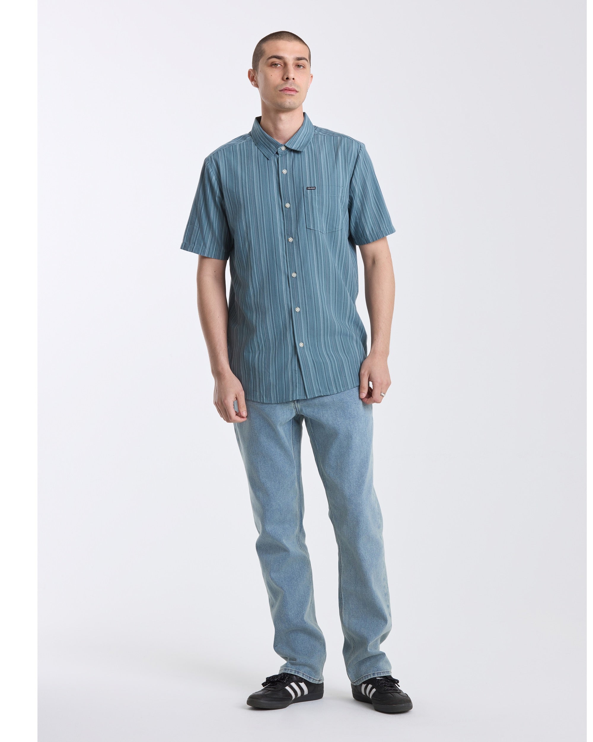 A person stands against a white background, wearing the Volcom Men's Jahstripe Short Sleeve Shirt, paired with light blue jeans and black sneakers with white stripes.