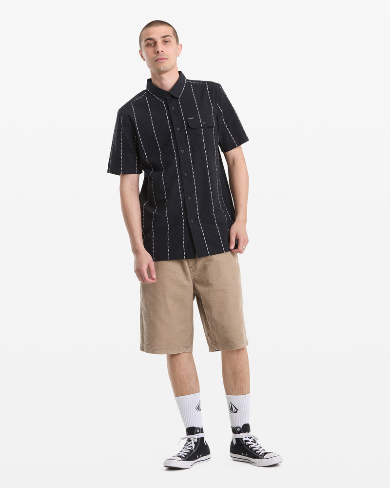 A man poses against a white backdrop in a black Volcom Men's Whitworth Short Sleeve shirt with white stripes, tan shorts, white crew socks, and black-and-white sneakers.