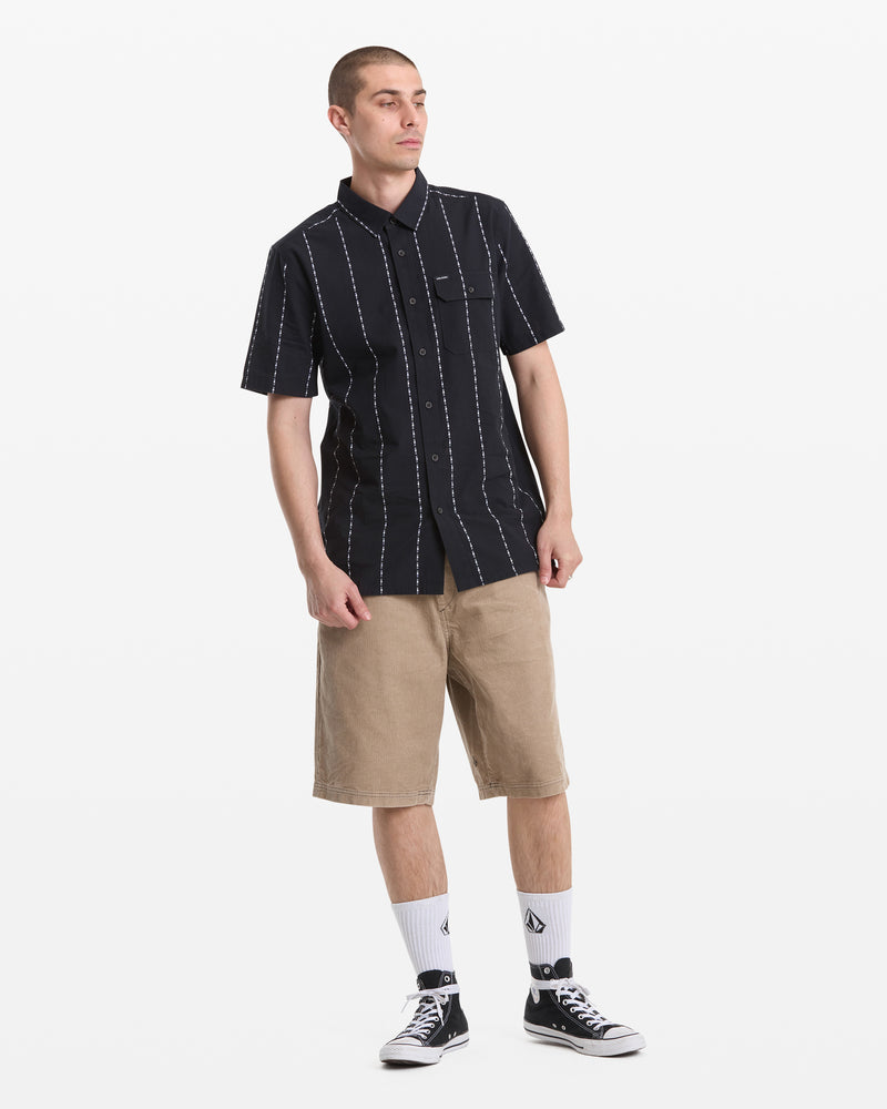 A person stands against a white background wearing the Volcom Men's Whitworth Short Sleeve shirt with white vertical stripes, tan knee-length shorts, logo white socks, and black-and-white sneakers.