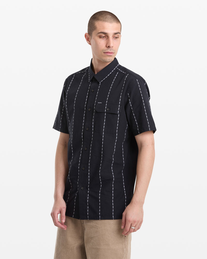 A man with short hair wears a Volcom Men's Whitworth Short Sleeve shirt in black with white vertical stripes and tan pants, standing against a plain white background.