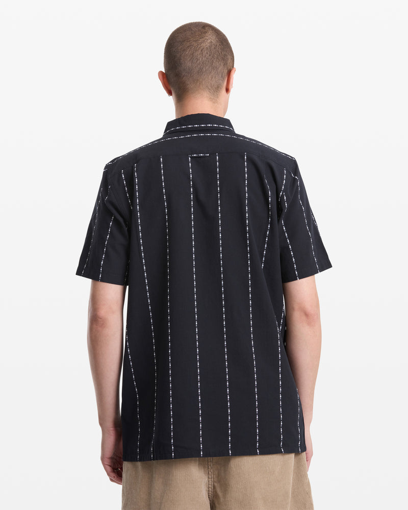 A person with short hair is seen from behind, wearing the Volcom Men's Whitworth Short Sleeve shirt in black with white vertical stripes and light brown shorts, standing against a plain white background.