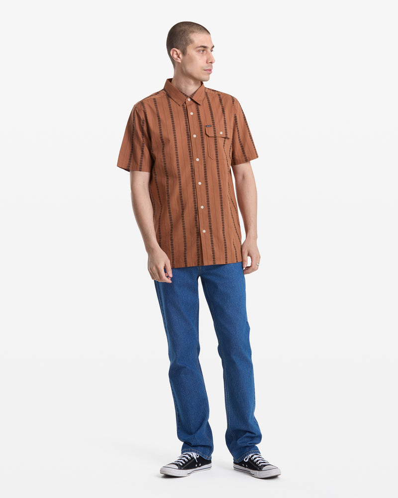 A person stands against a white background wearing the Volcom Men's Whitworth Short Sleeve, a brown patterned button-up shirt with jacquard stripes, paired with blue jeans and black sneakers. They look slightly to the side with one hand near their pocket.