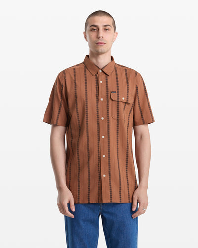 A man with short hair wears the Volcom Men's Whitworth Short Sleeve, a brown 100% cotton shirt with vertical black stripes, and blue jeans, standing against a plain white background.