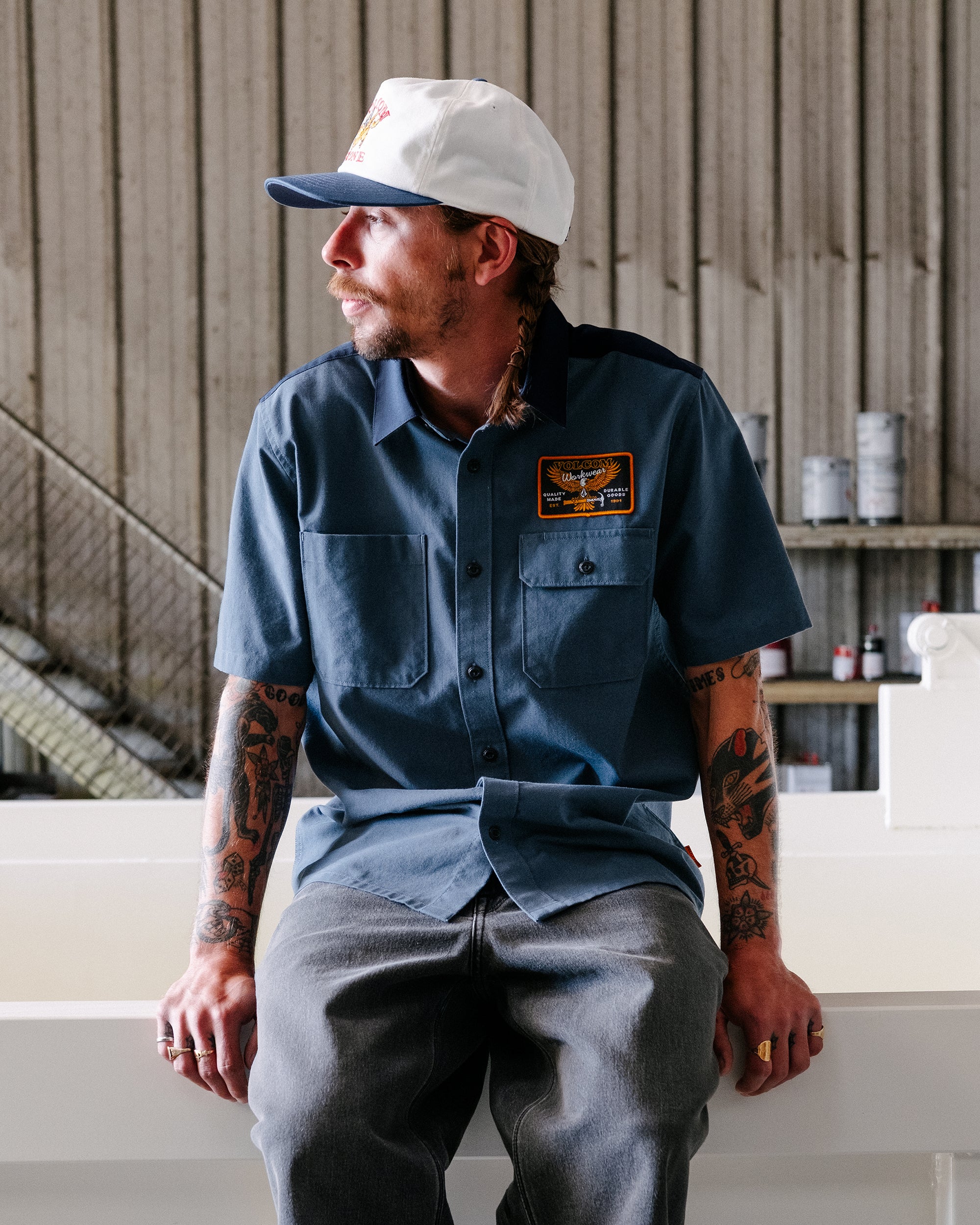 A tattooed man wears the Volcom Men's Slab City Short Sleeve Tee, gray jeans, and a white cap while sitting indoors beside an industrial-style wall with shelves and metal stairs, looking to the left.
