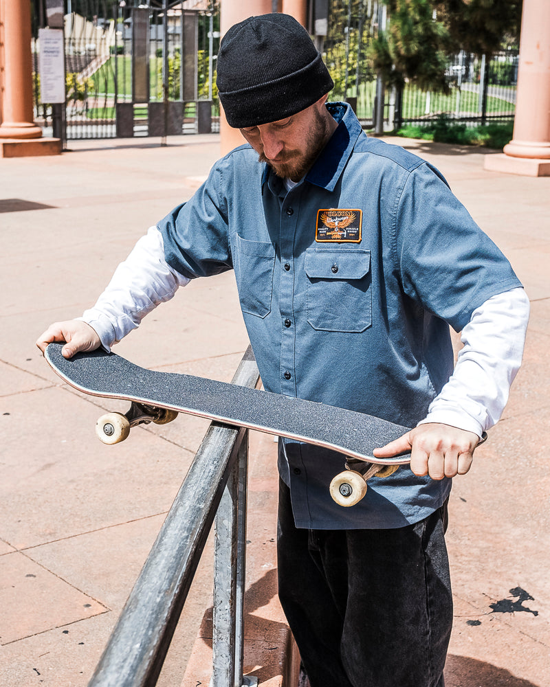 A man in a Volcom Men's Slab City Short Sleeve Tee and beanie holds a skateboard by a metal rail outdoors, looking ready to attempt a trick at the skate park.