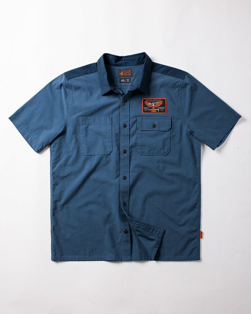 The Volcom Men's Slab City Short Sleeve Tee is shown in blue with two chest pockets and a black and orange patch above the right pocket, made from a durable cotton-poly blend, laid flat on a white background.
